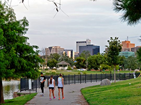 People walking on sidewalk near pond with city in background